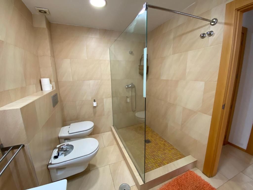 Baño con ducha con mampara de cristal, dos inodoros y paredes de azulejos beige.