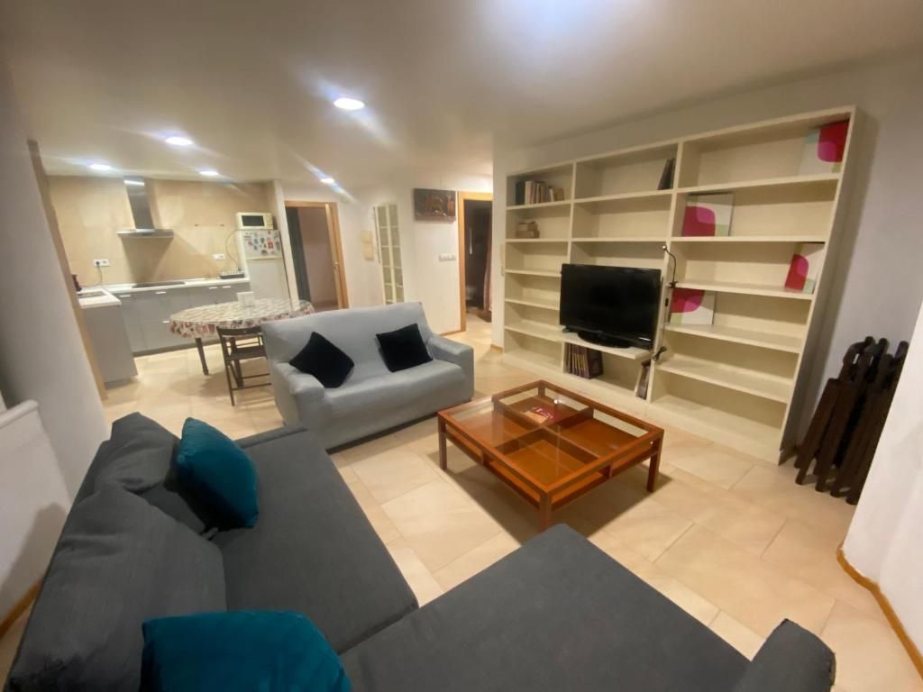 Sala de estar con sofás grises, estanterías empotradas, televisor y mesa de centro. La cocina se ve al fondo.