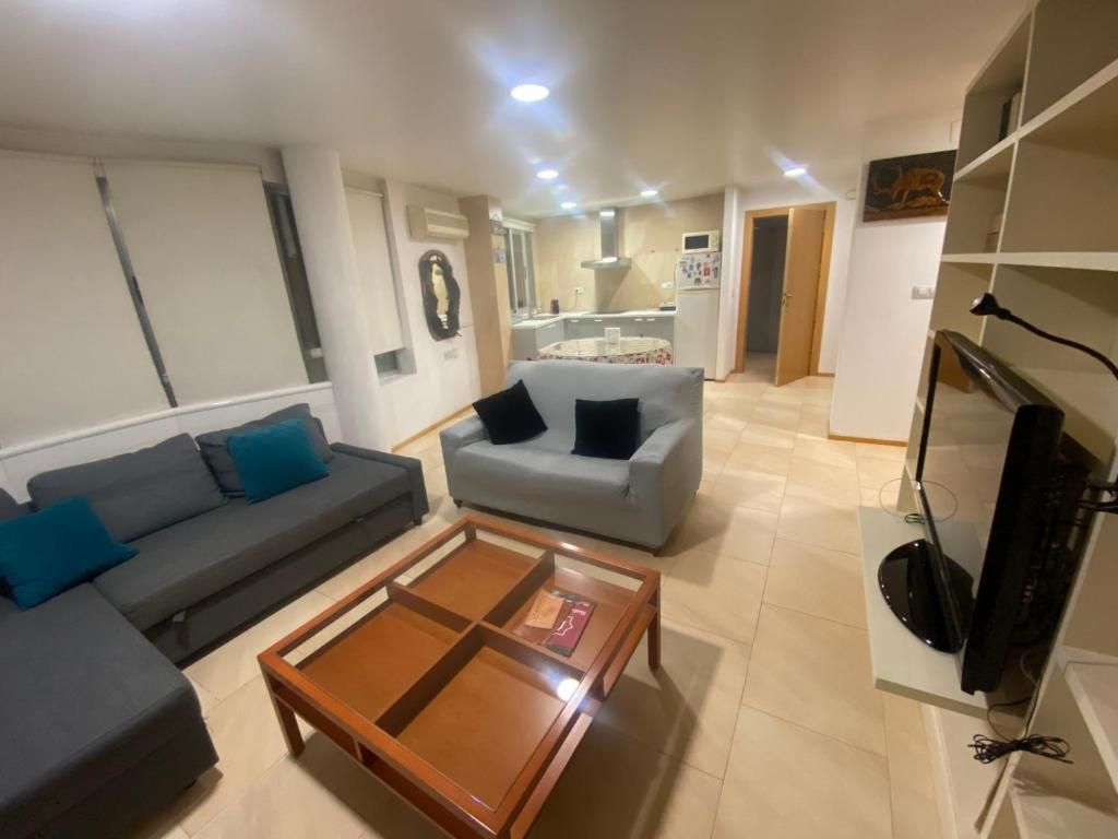 Sala de estar con sofás grises, mesa de centro de madera y cocina abierta visible al fondo.