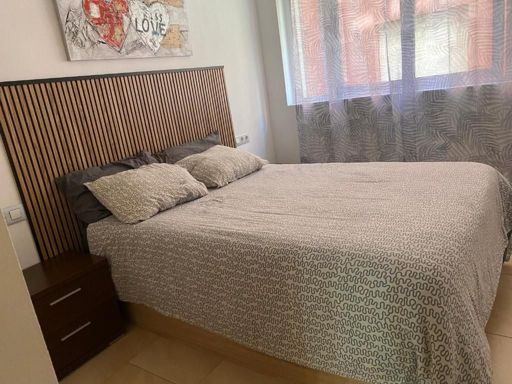 Dormitorio con cama, funda estampada, almohadas y cabecero de madera. Mesilla de noche y cuadro sobre la cama.