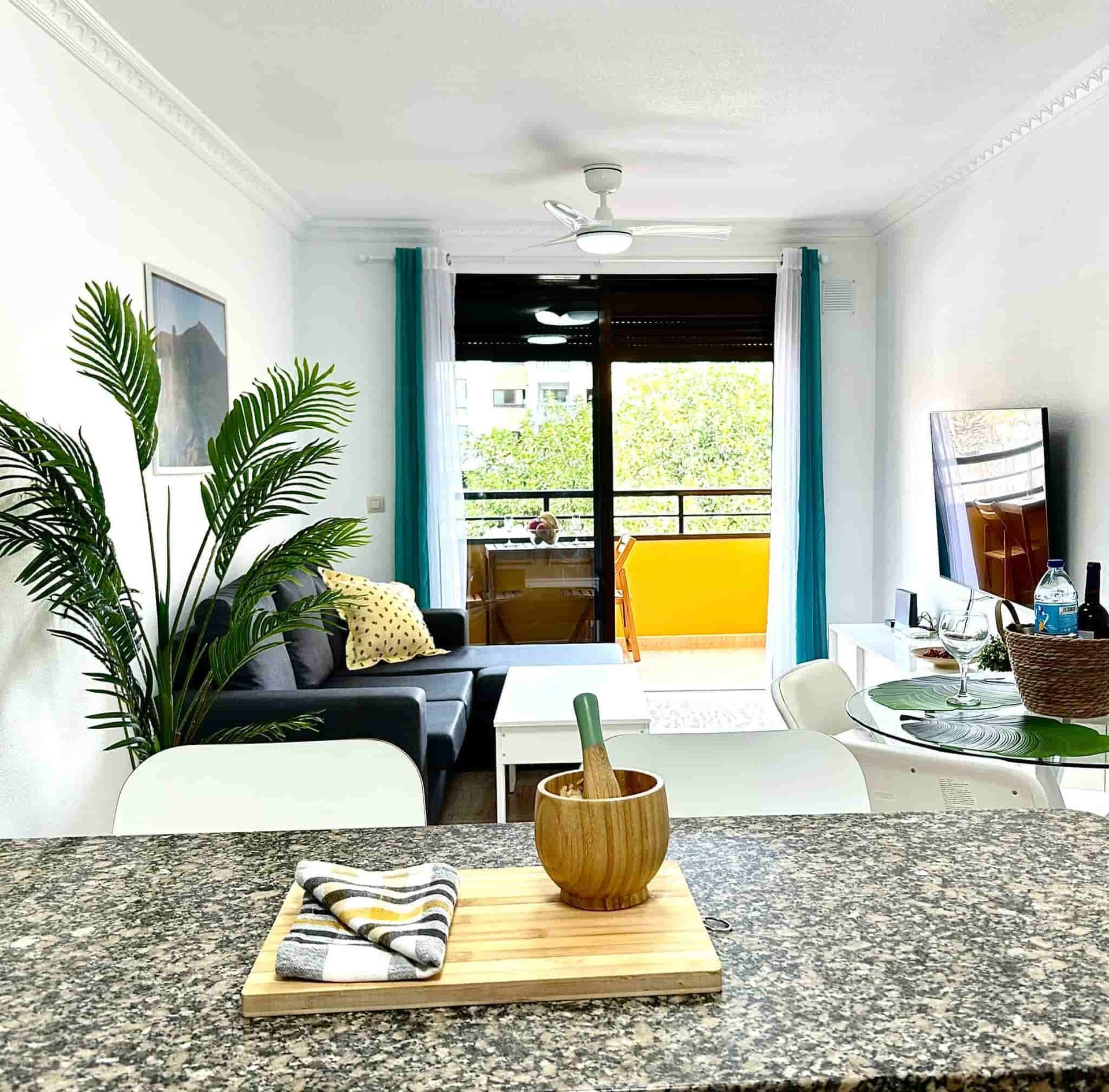 Sala de estar con acceso al balcón; sofá gris, cortinas verde azulado, muebles blancos y una encimera de cocina en primer plano.