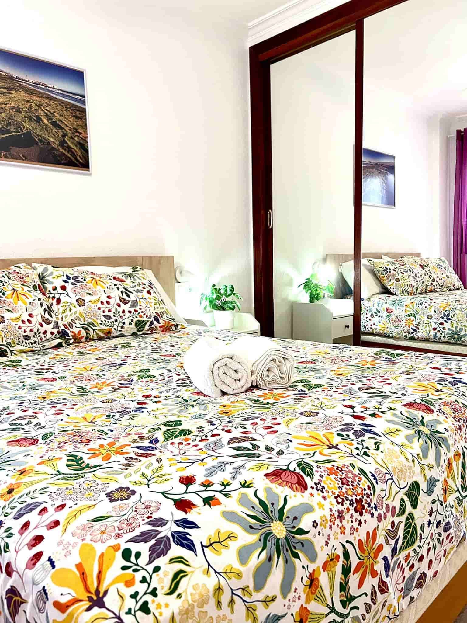 Dormitorio con ropa de cama floral, paredes blancas, espejo grande que refleja la cama y obras de arte.