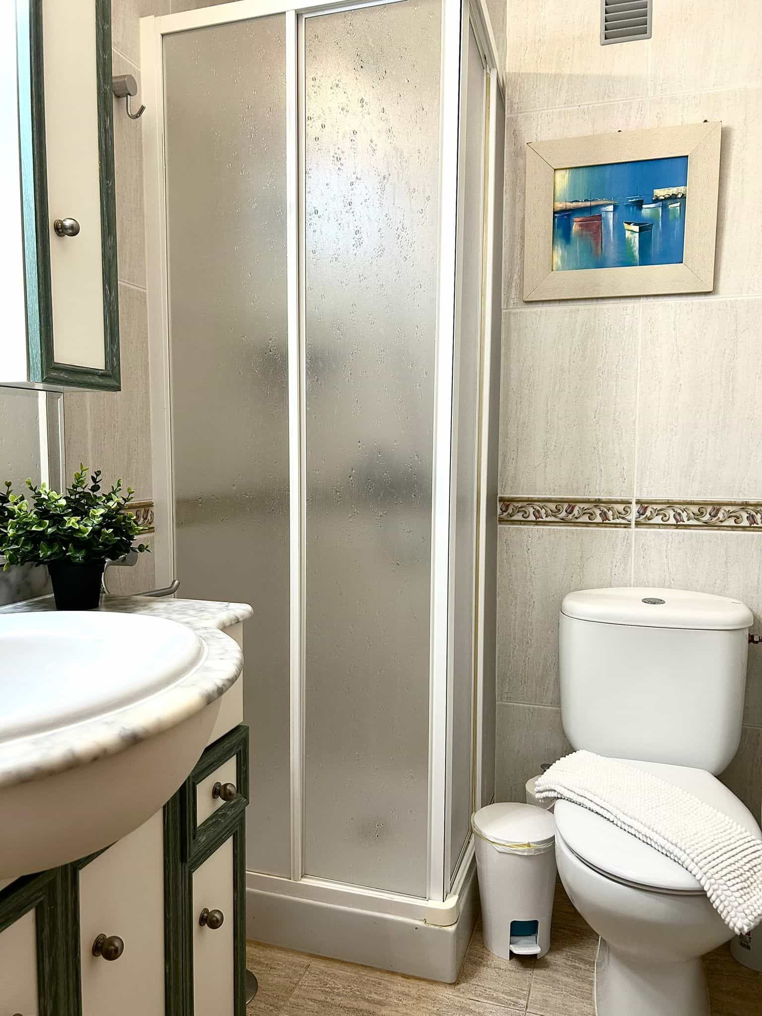 Baño con ducha de esquina, inodoro, lavabo y obras de arte en una pared de azulejos.