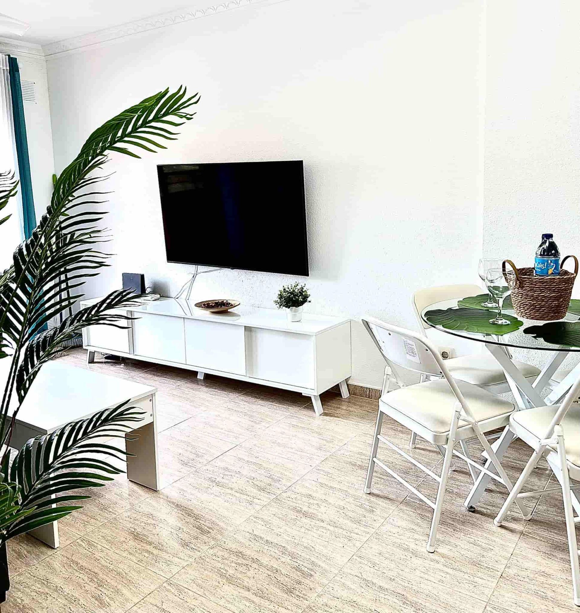 Sala de estar con muebles blancos, TV y planta artificial.