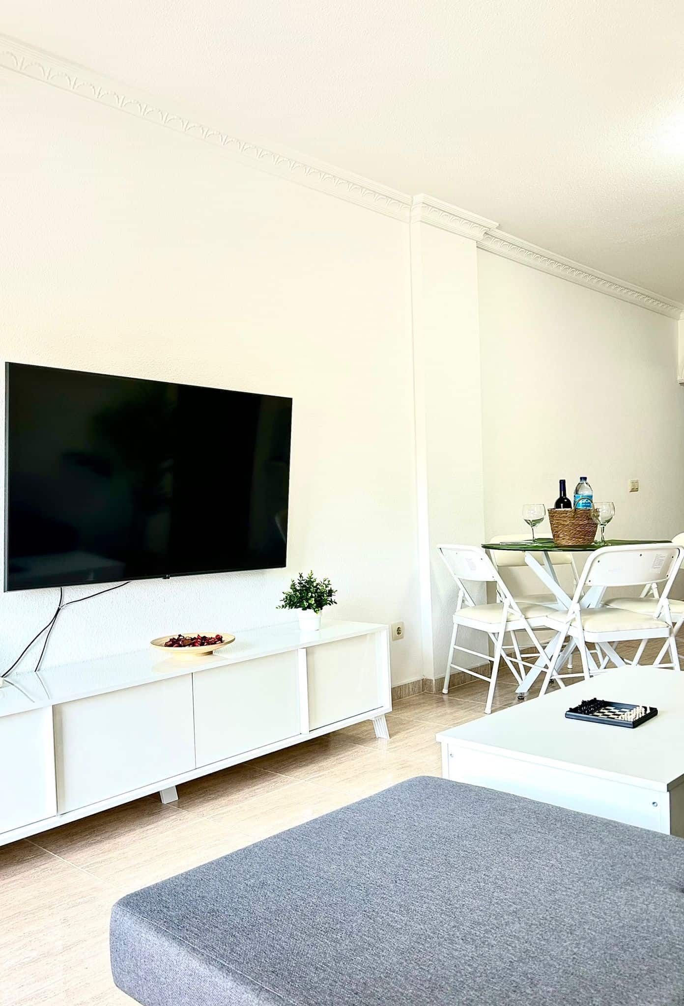 Moderna sala de estar con un televisor grande, muebles blancos y una pequeña mesa de comedor.