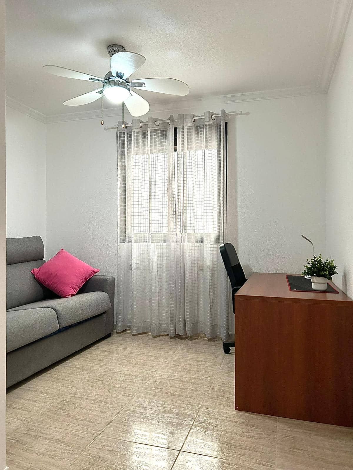 Habitación con sofá gris, escritorio, ventana, ventilador de techo y alfombra beige.