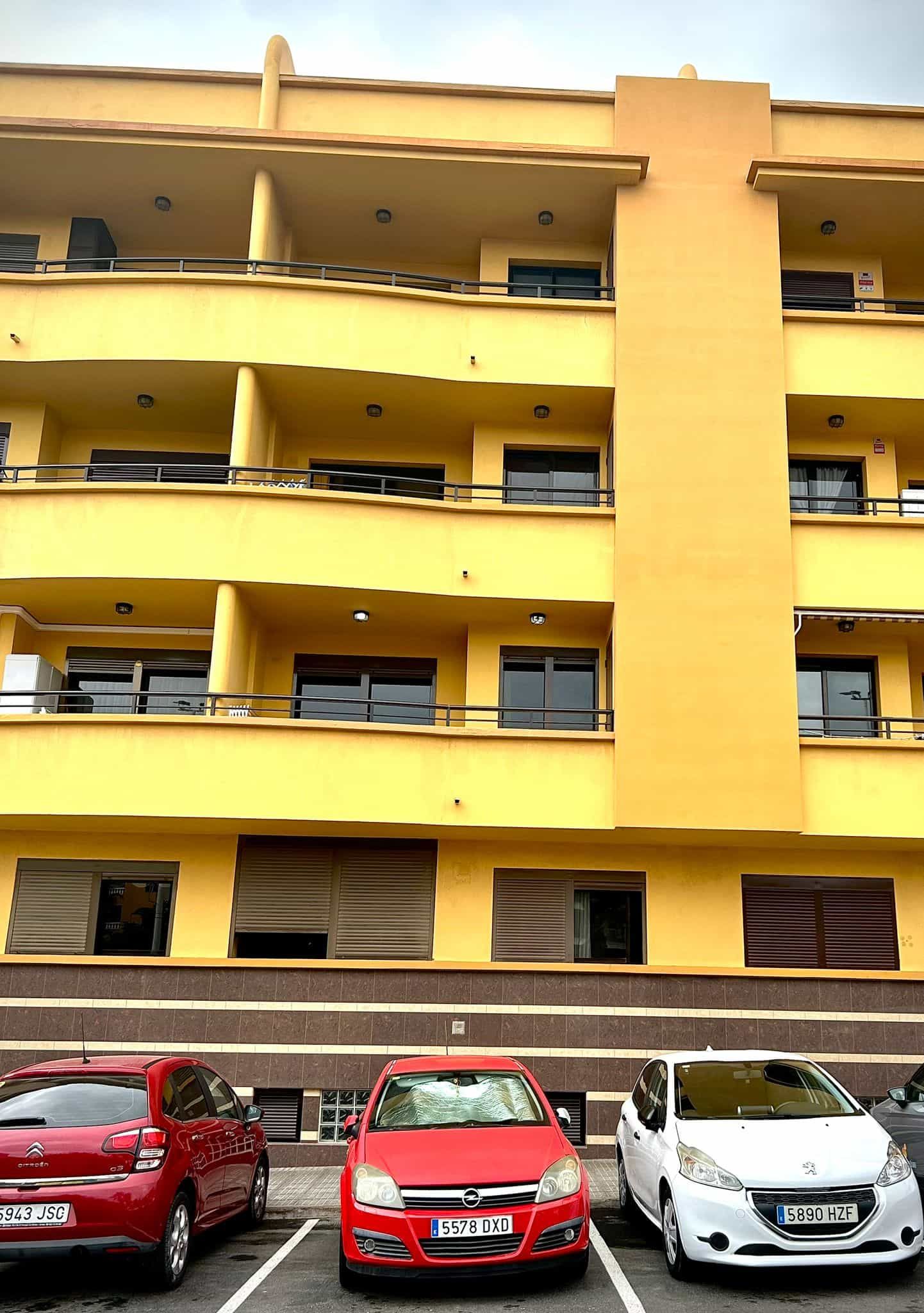 Edificio de apartamentos amarillo con balcones, tres coches aparcados delante.