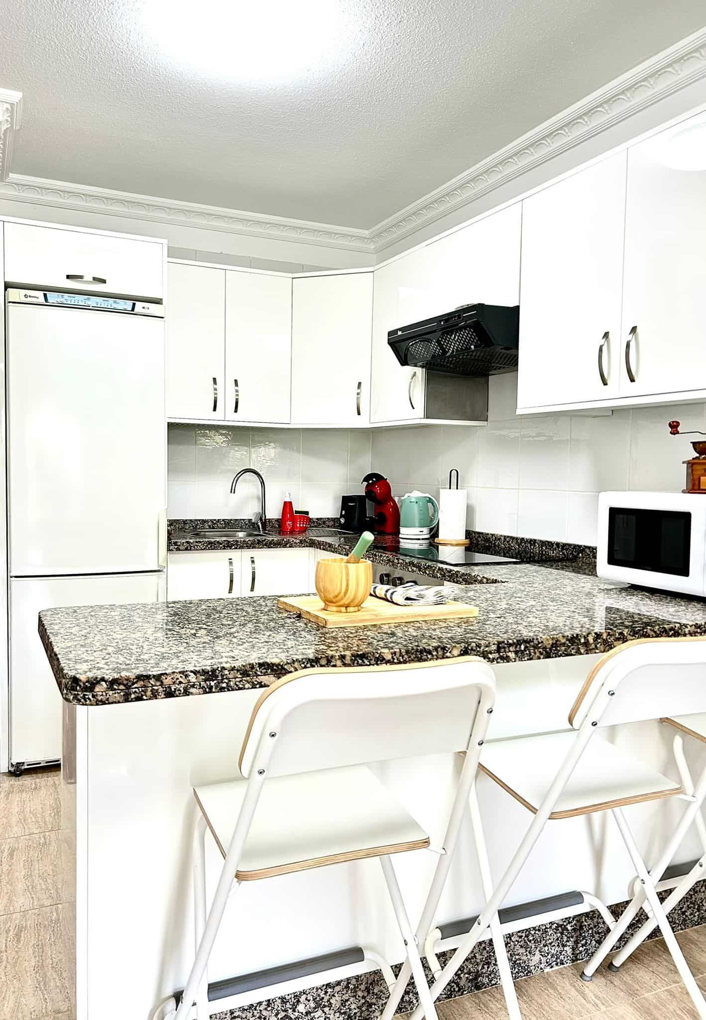 Cocina blanca con barra de desayuno, gabinetes blancos, encimera de granito y dos sillas plegables blancas.
