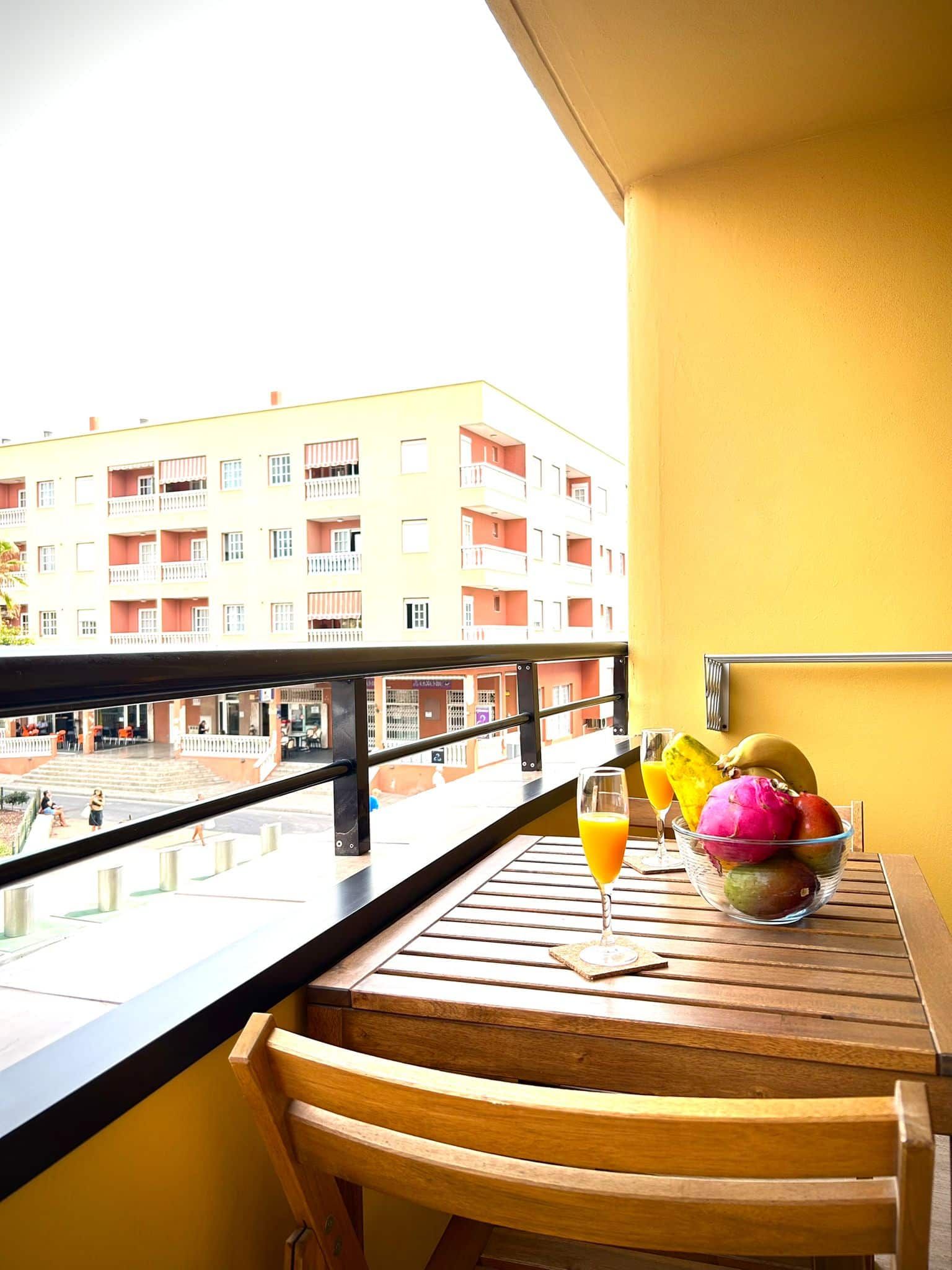 Balcón con mesa, silla, fruta, bebida y vista a un edificio amarillo.