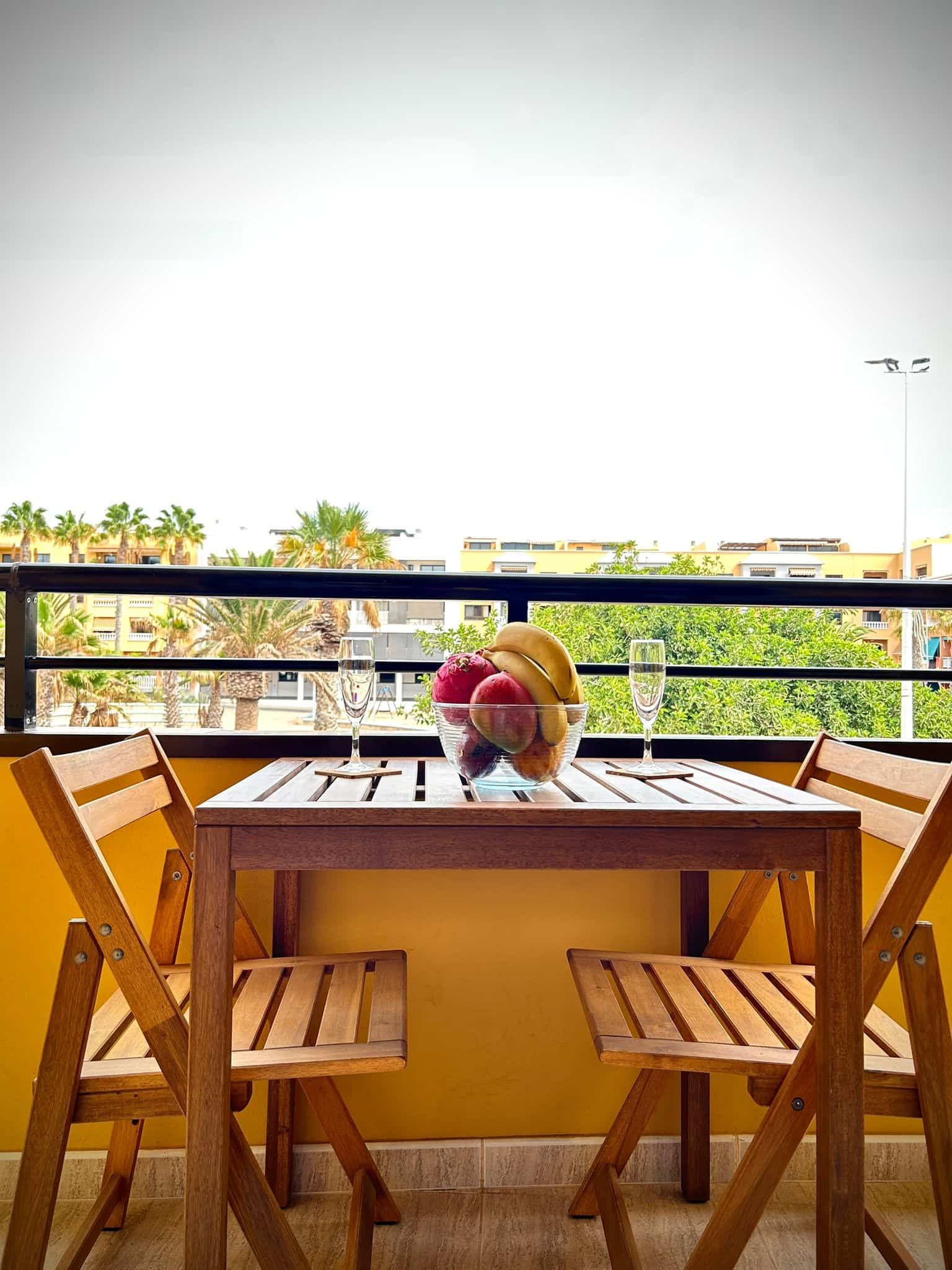 Mesa y sillas de madera en un balcón con frutero. Vistas a árboles y edificios.