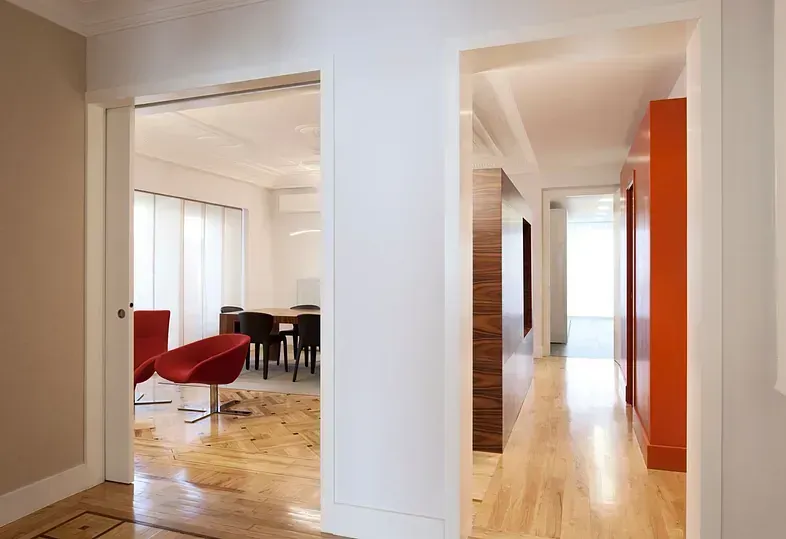 Pasillo con suelo de madera que da acceso al comedor. Sillas rojas, armarios naranjas y paredes blancas.