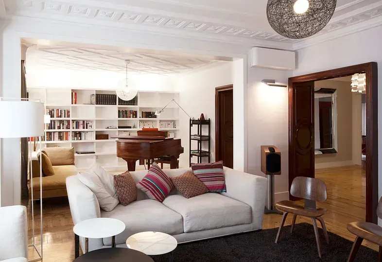 Sala de estar: sofá blanco, mesas de café, piano, estantería, alfombra oscura, piso de madera.