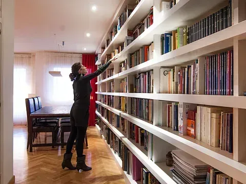 Mujer alcanzando un libro en una estantería larga en una habitación bien iluminada con una mesa de comedor.