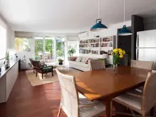 Luminosa sala de estar con comedor. Paredes blancas, suelos de madera y amplios ventanales. Amueblada con sofá, mesa, sillas y librería.