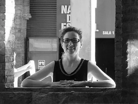 Una mujer con gafas sonríe, enmarcada por una abertura de ladrillo. Edificio y letrero al fondo.