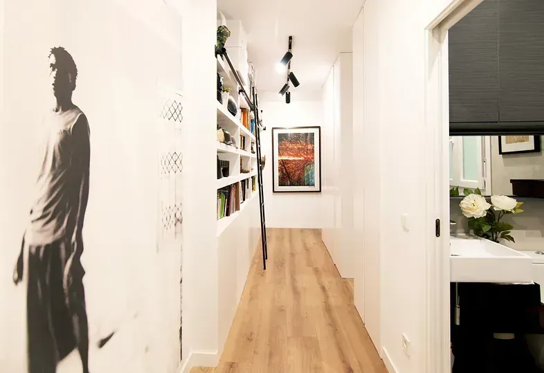 Pasillo estrecho de color blanco con estanterías empotradas, una obra de arte enmarcada y una gran figura en escala de grises.