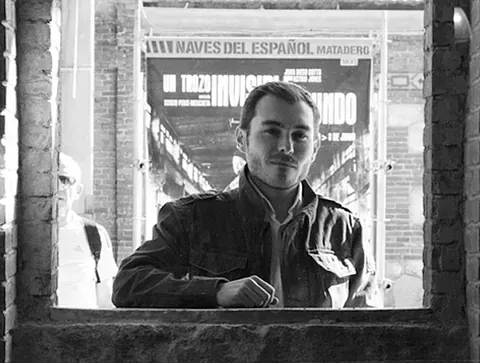 Hombre con chaqueta enmarcado en una ventana, mirando a la cámara; edificio en el fondo, en blanco y negro.