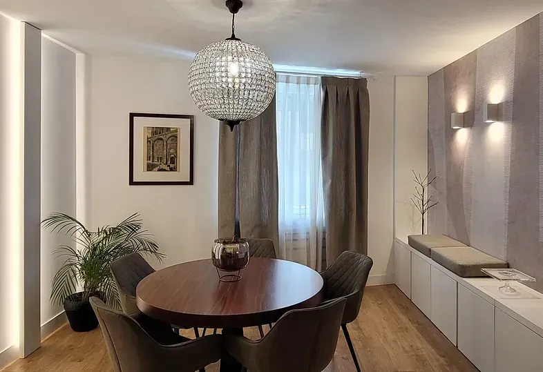 Comedor con mesa redonda de madera, sillas grises, araña de cristal y cortinas beige.
