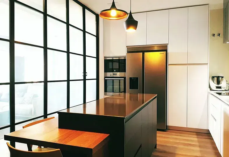 Cocina moderna con isla, pared de ventanas esmeriladas, electrodomésticos de acero inoxidable y luces colgantes.