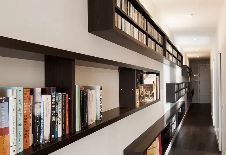Pasillo largo con estanterías oscuras empotradas que exhiben libros y CD. Paredes blancas y suelos de madera oscura.