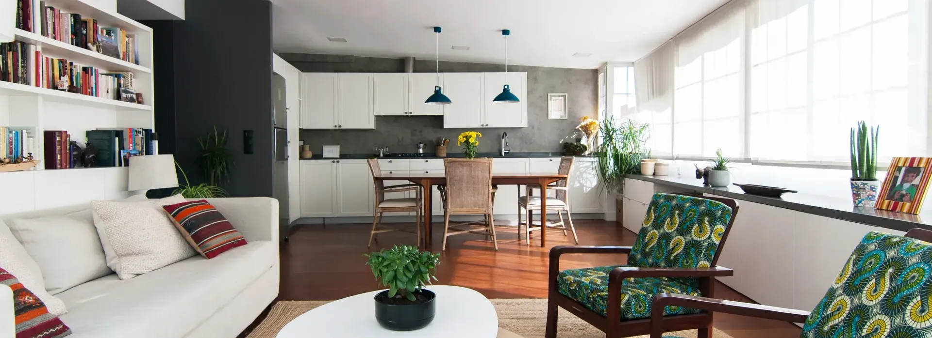 Una sala de estar moderna con un sofá blanco, sillas con tela estampada verde y una cocina al fondo.