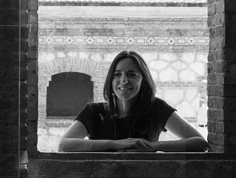 Mujer sonriendo, enmarcada por una ventana de ladrillo, con los brazos en la cornisa, edificio de fondo.