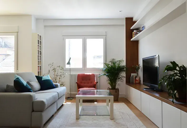 Sala de estar con paredes blancas, sofá gris, TV y un sillón naranja junto a una ventana.