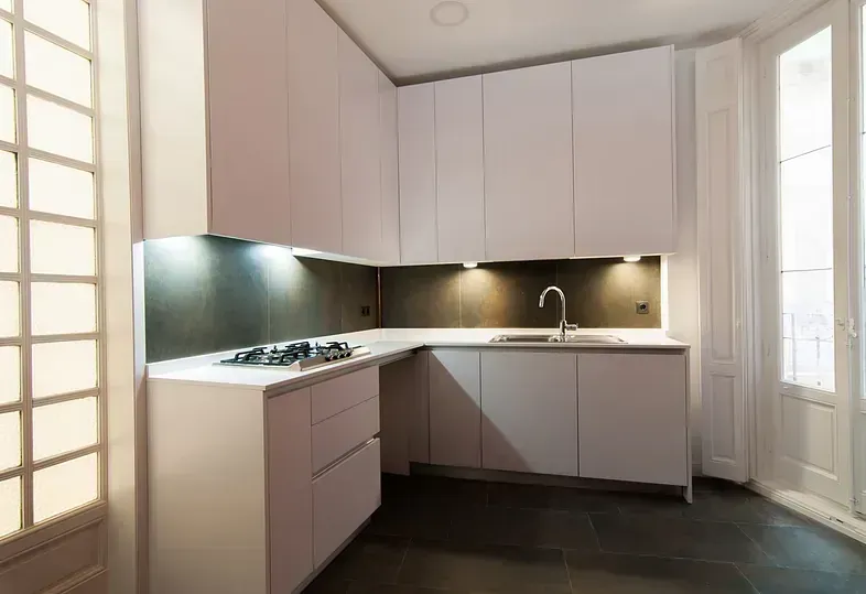 Cocina moderna blanca con gabinetes, fregadero y estufa. Luz natural por la ventana.