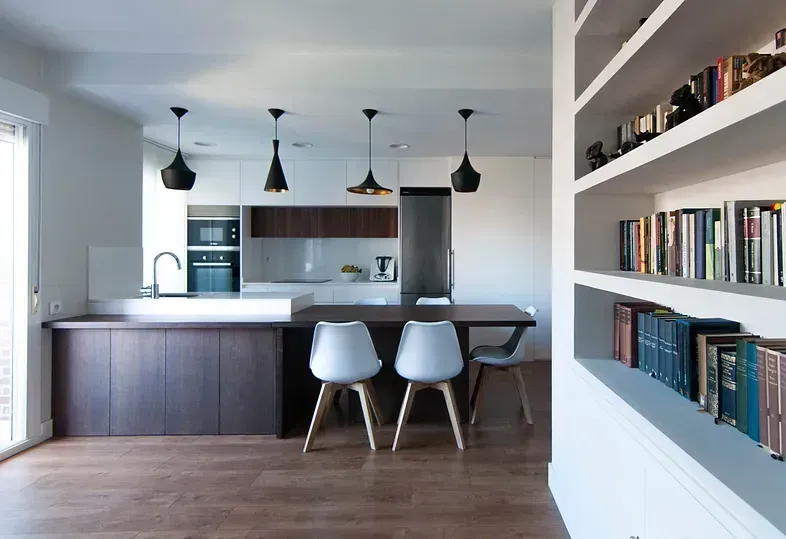 Cocina moderna con gabinetes de madera oscura, paredes blancas y lámparas colgantes. Estanterías a la derecha.