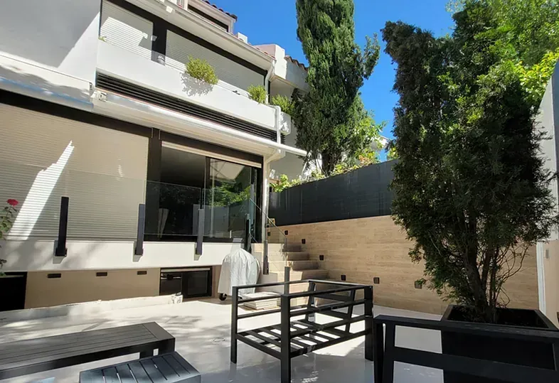 Exterior de edificio moderno con patio, escalones, árboles y asientos; luz solar brillante.