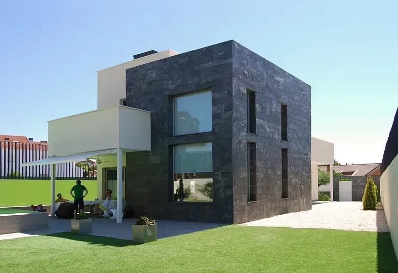 Casa moderna con fachada de piedra negra, detalles blancos, césped verde y gente en el patio.