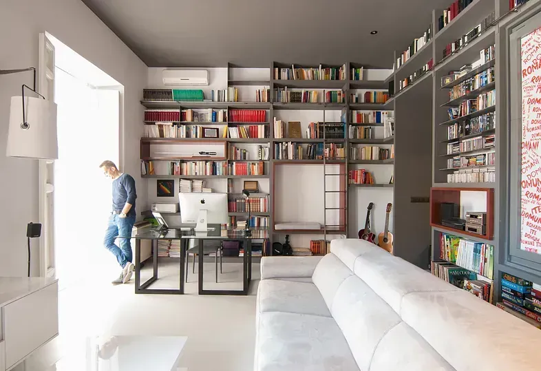 Hombre de pie cerca de una puerta abierta en una oficina en casa con estanterías, escritorio, sofá y guitarras.