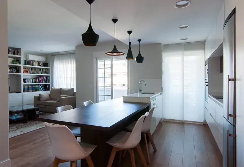 Moderna cocina abierta y sala de estar con mesa de isla oscura, sillas blancas y luces colgantes negras.