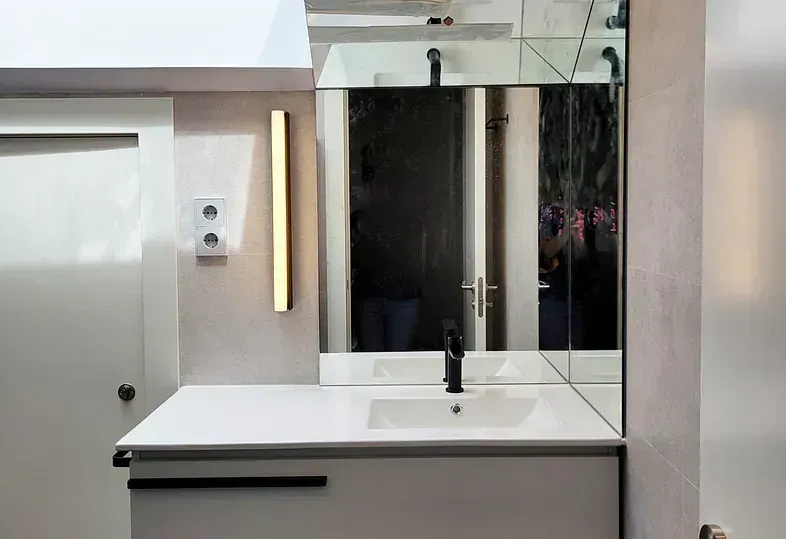 Baño moderno con tocador blanco, grifería negra, espejo y aplique de pared vertical dorado.