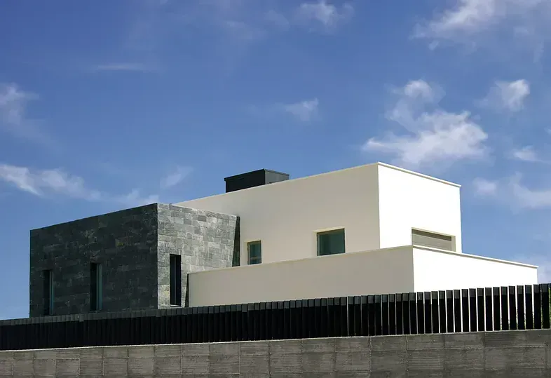 Casa moderna blanca y gris con valla negra contra un cielo azul.
