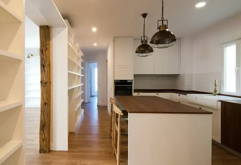 Cocina con gabinetes blancos, isla de madera, luces colgantes industriales y estanterías empotradas.
