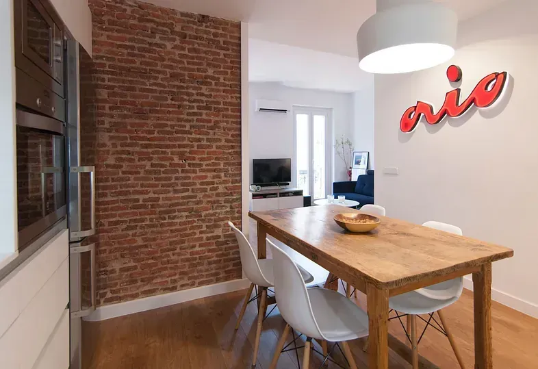 Comedor con mesa de madera, sillas blancas, pared de ladrillo visto y cartel de “rio”.