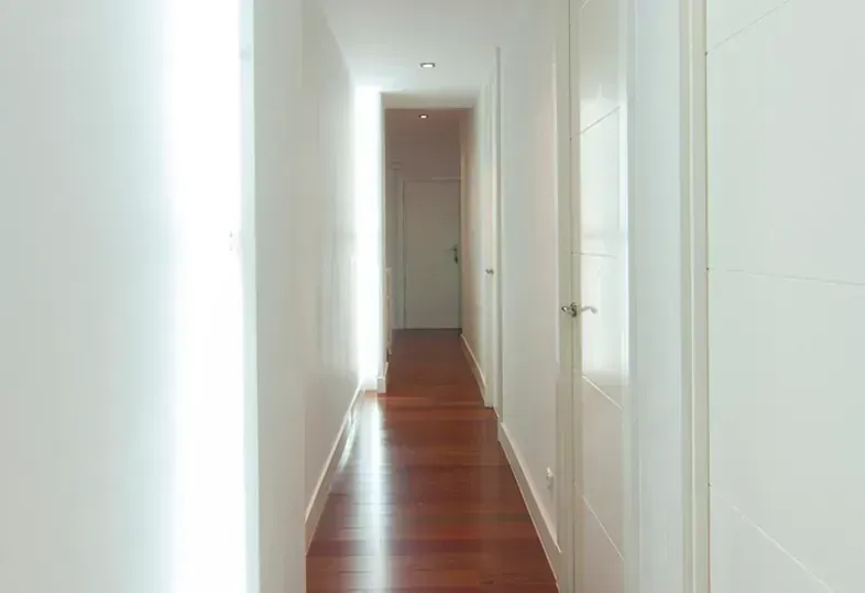 Pasillo largo y estrecho con suelo de madera, paredes y puertas blancas. La luz del sol entra por la izquierda.