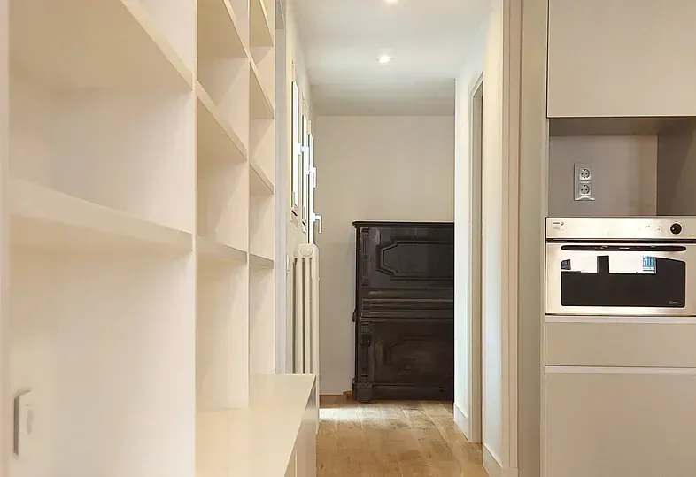 Pasillo estrecho con estanterías empotradas a un lado y un horno al otro. Un piano al fondo.