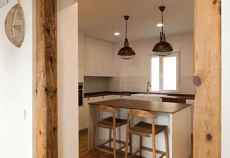 Cocina con gabinetes blancos, isla de madera y luces colgantes rústicas; vista a través de la puerta de madera.