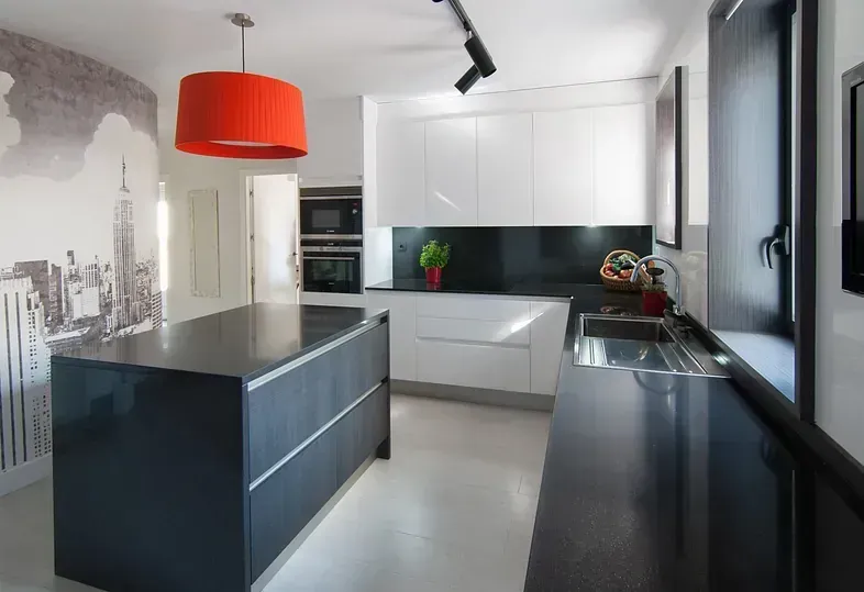 Cocina moderna con isla y encimeras negras, gabinetes blancos y una lámpara colgante naranja.