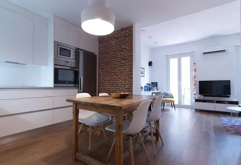 Cocina abierta y comedor con mesa de madera y sillas blancas, pared de ladrillo y pisos de madera clara.