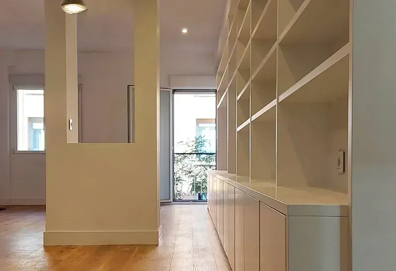 Pasillo interior con estanterías y armarios empotrados en color blanco, que conduce a un balcón con vegetación.