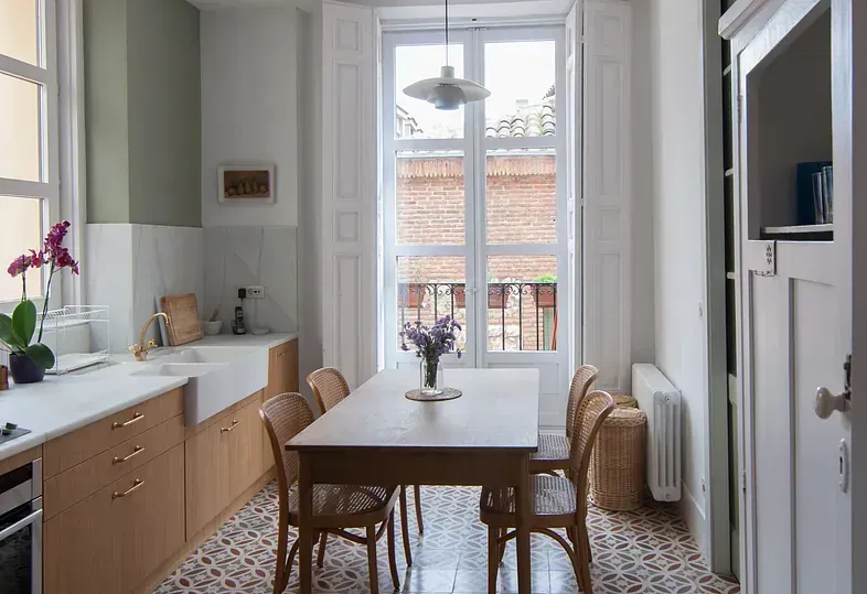 Cocina con gabinetes de madera clara, mesa de comedor, piso estampado y una ventana que da a un balcón.