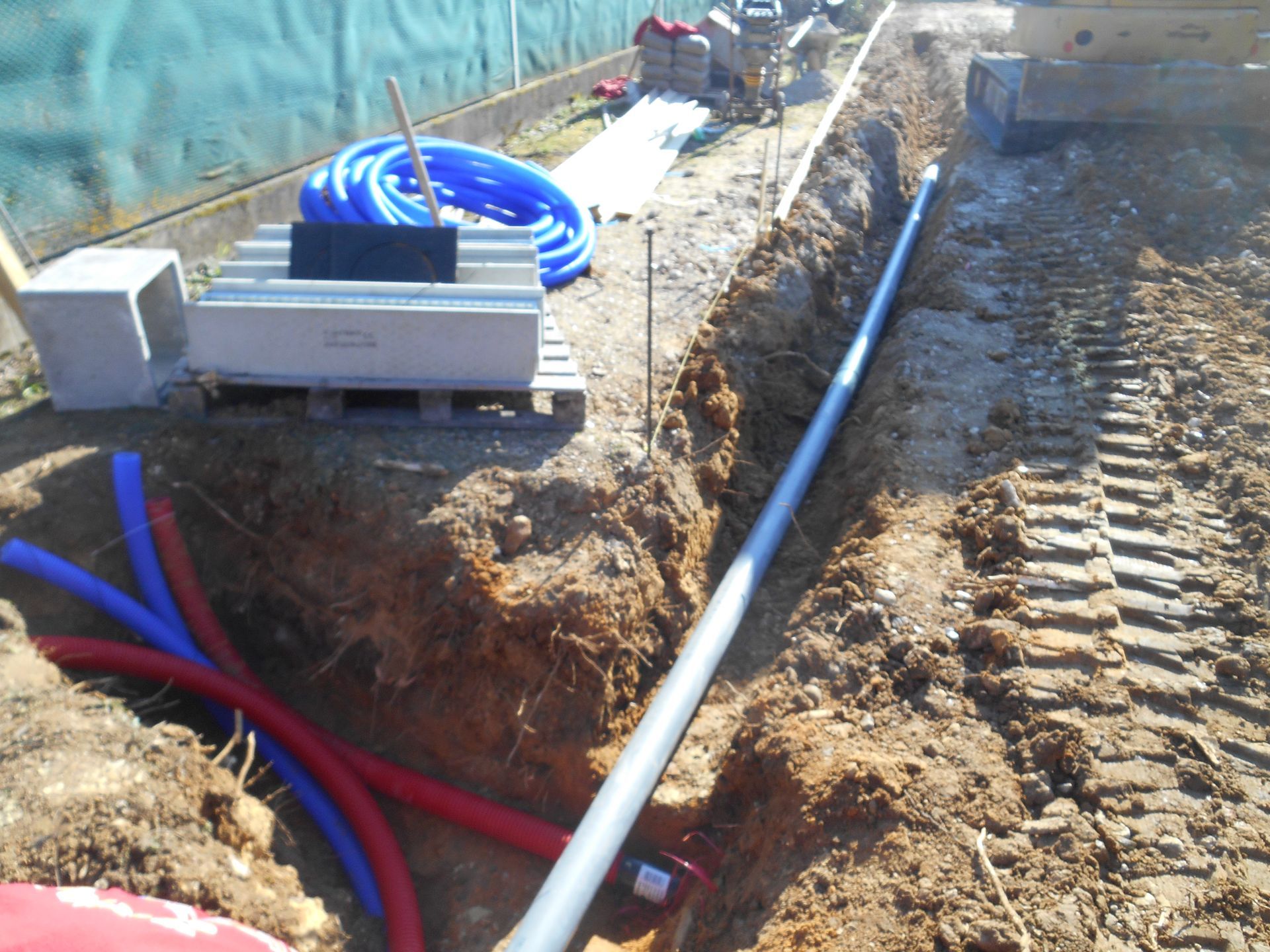 Tranchée technique avec des tuyaux bleus et rouges et une machine blanche le long d'un chantier d'excavation.