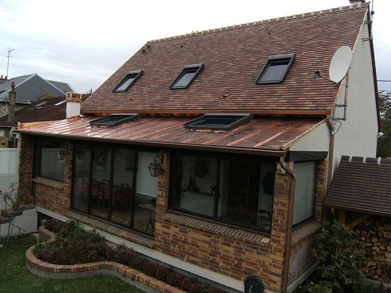 Maison en briques avec toit de tuiles rouges, puits de lumière et grandes fenêtres en façade sous un ciel nuageux