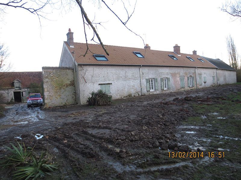 Cour boueuse jouxtant une longue ferme en pierre au toit rouge.