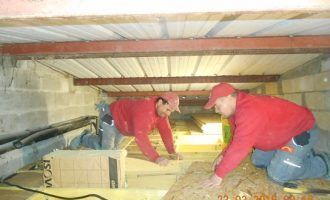 Deux ouvriers vêtus de rouge sont accroupis dans un vide sanitaire bas, en train d'inspecter ou d'installer du matériel sous un plancher.