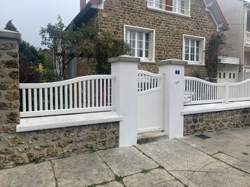 Maison en pierre avec clôture et portail blancs, donnant sur un trottoir, avec un petit panneau bleu sur le portail.