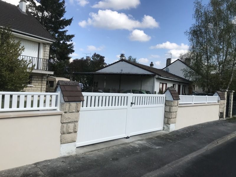 Clôture et portail blancs le long d'une rue résidentielle bordée de maisons et d'arbres sous un ciel bleu.